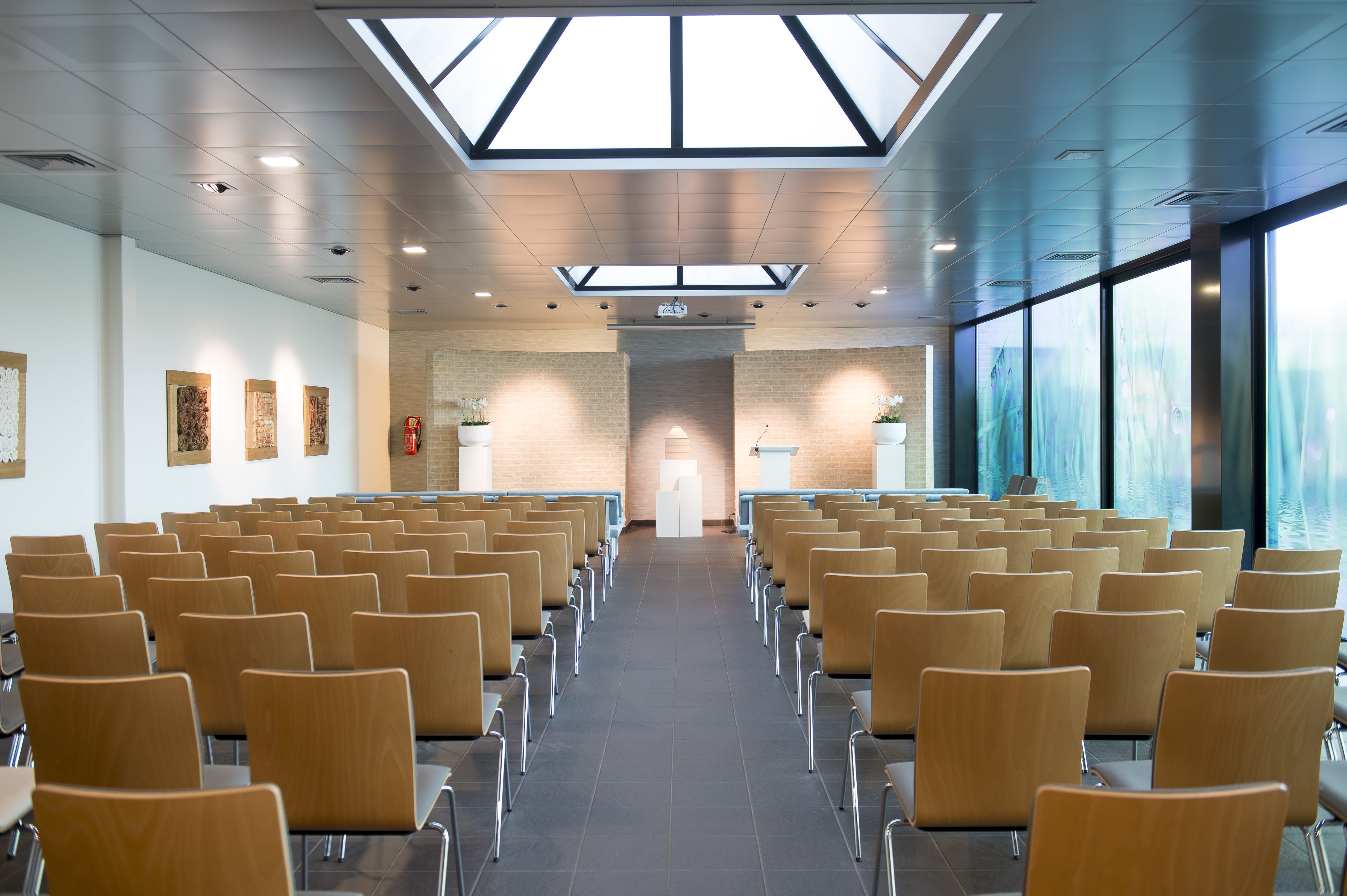 Aula met glazen puntdak, grote ramen en houten stoelen op meerdere rijen
